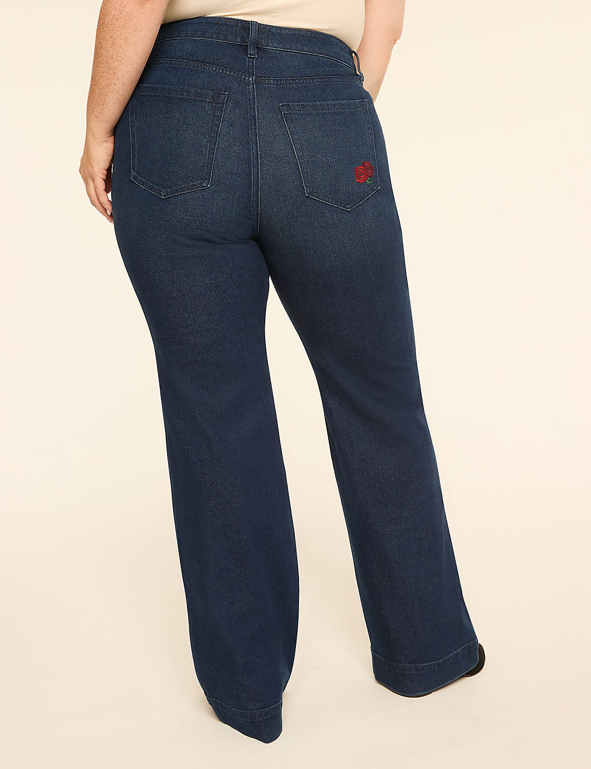 A person wearing dark wash flared jeans with a red embroidered logo on the back pocket stands against a plain, light-colored background. Product Image 2