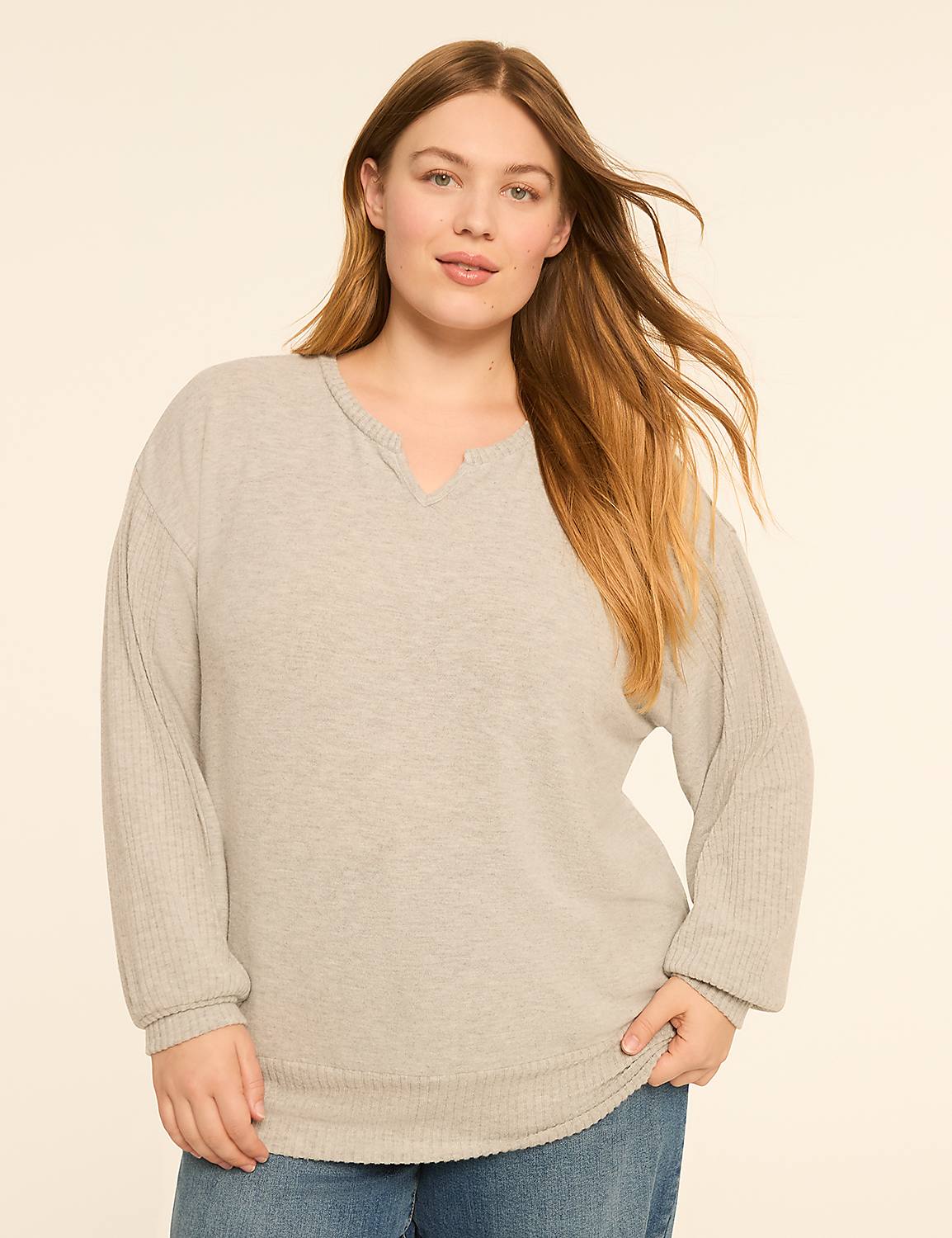 A woman with long, blonde hair stands in the foreground wearing a light gray, long-sleeved sweater and blue jeans, against a plain, light beige background. Product Image 1