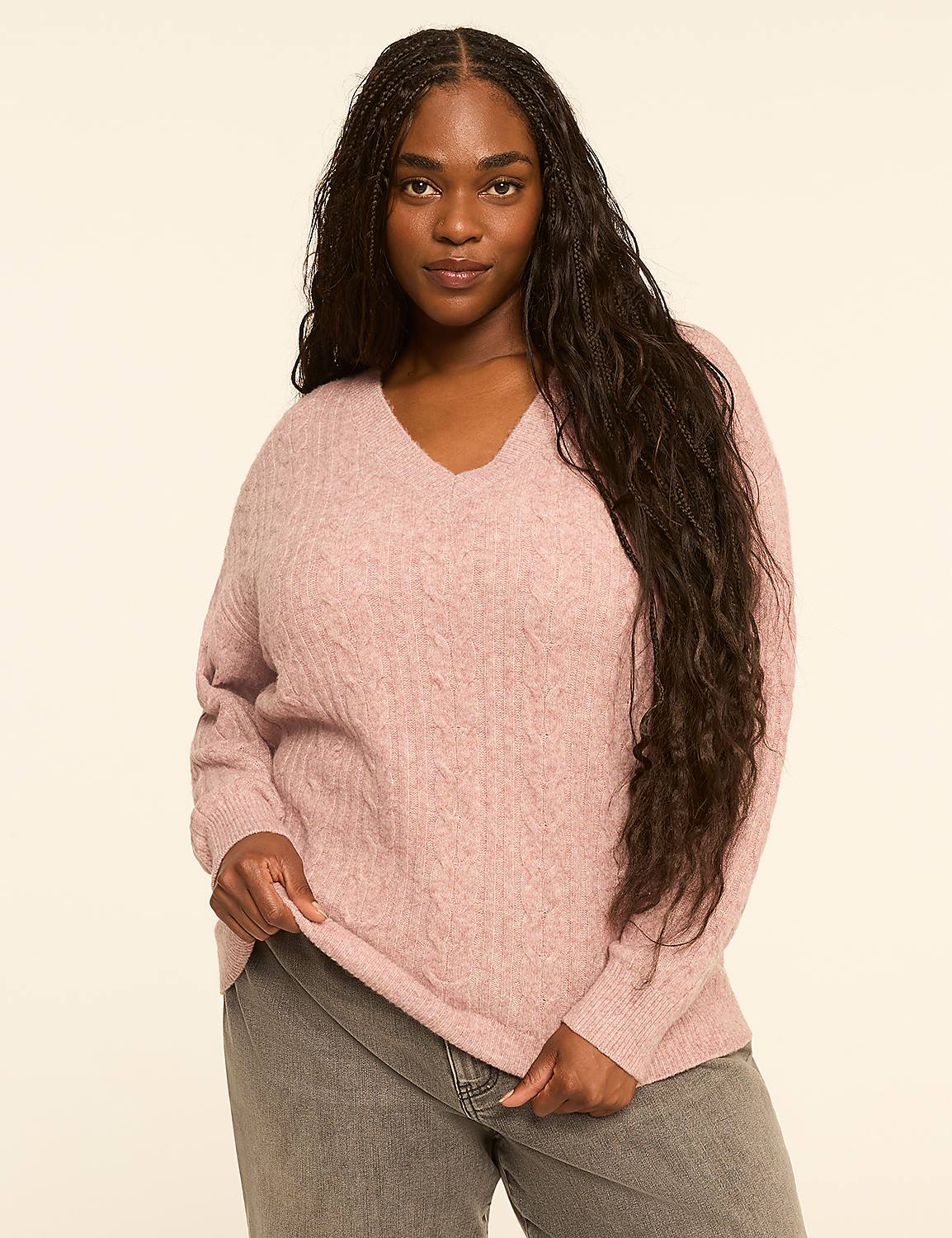 A woman with long, dark hair stands in the foreground wearing a pink cable-knit sweater and gray jeans against a plain, light-colored background. Product Image 1