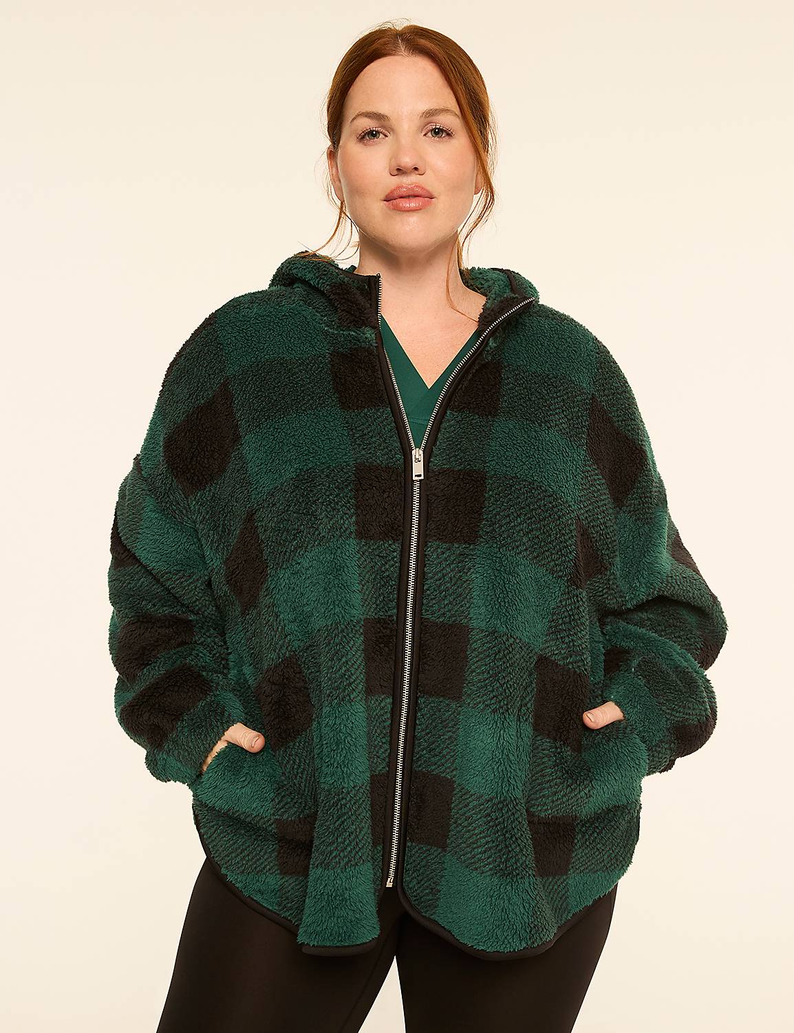 A woman with auburn hair stands in the foreground wearing a green and black buffalo plaid fuzzy zip-up hoodie over a green v-neck shirt, against a plain cream-colored background. Product Image 1