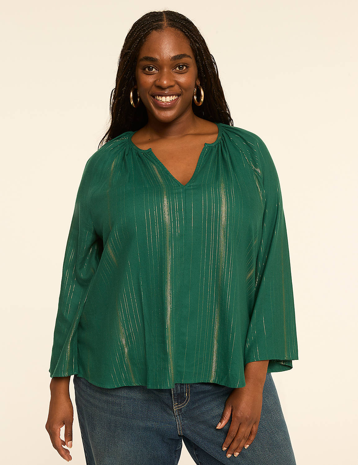A smiling woman with dark hair and hoop earrings, wearing a green, metallic-striped blouse and blue jeans, stands against a plain, light-colored background. Product Image 1
