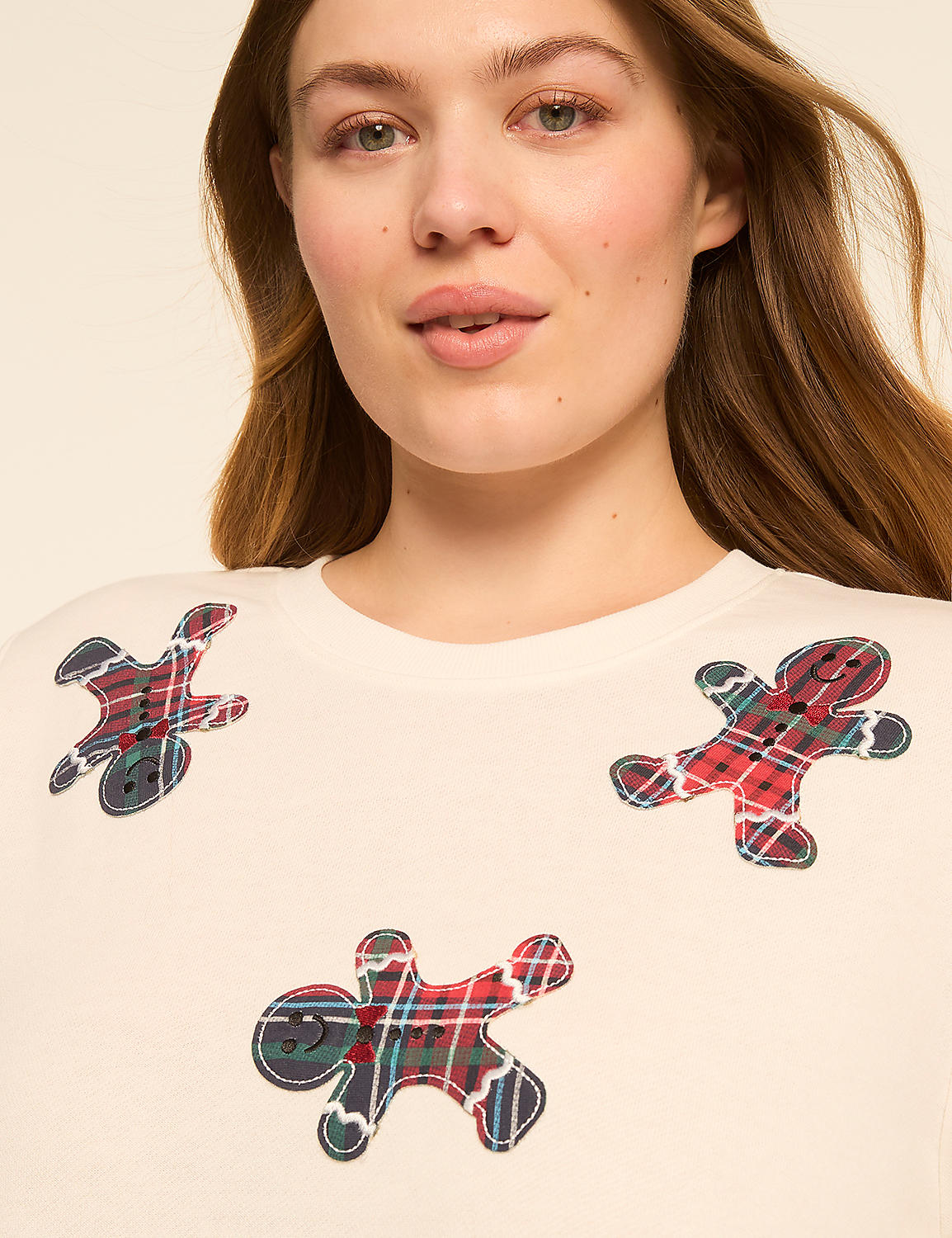 A woman with light skin and brown hair smiles gently, her cream-colored sweater adorned with three gingerbread man appliques in plaid fabric against a plain, light background. Product Image 4