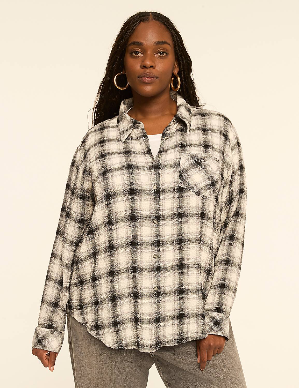 A woman with dark hair and hoop earrings poses in a cream and black plaid shirt and grey jeans against a plain, light-colored background. Product Image 1