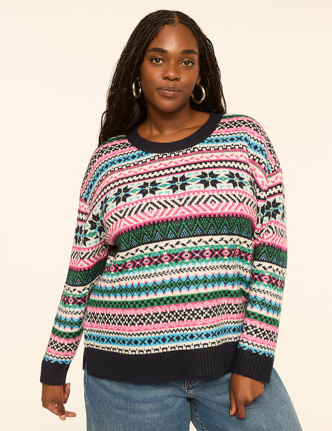 A woman with braided hair and hoop earrings poses in a colorful, patterned sweater and blue jeans against a plain, light-colored background. Product Image 1