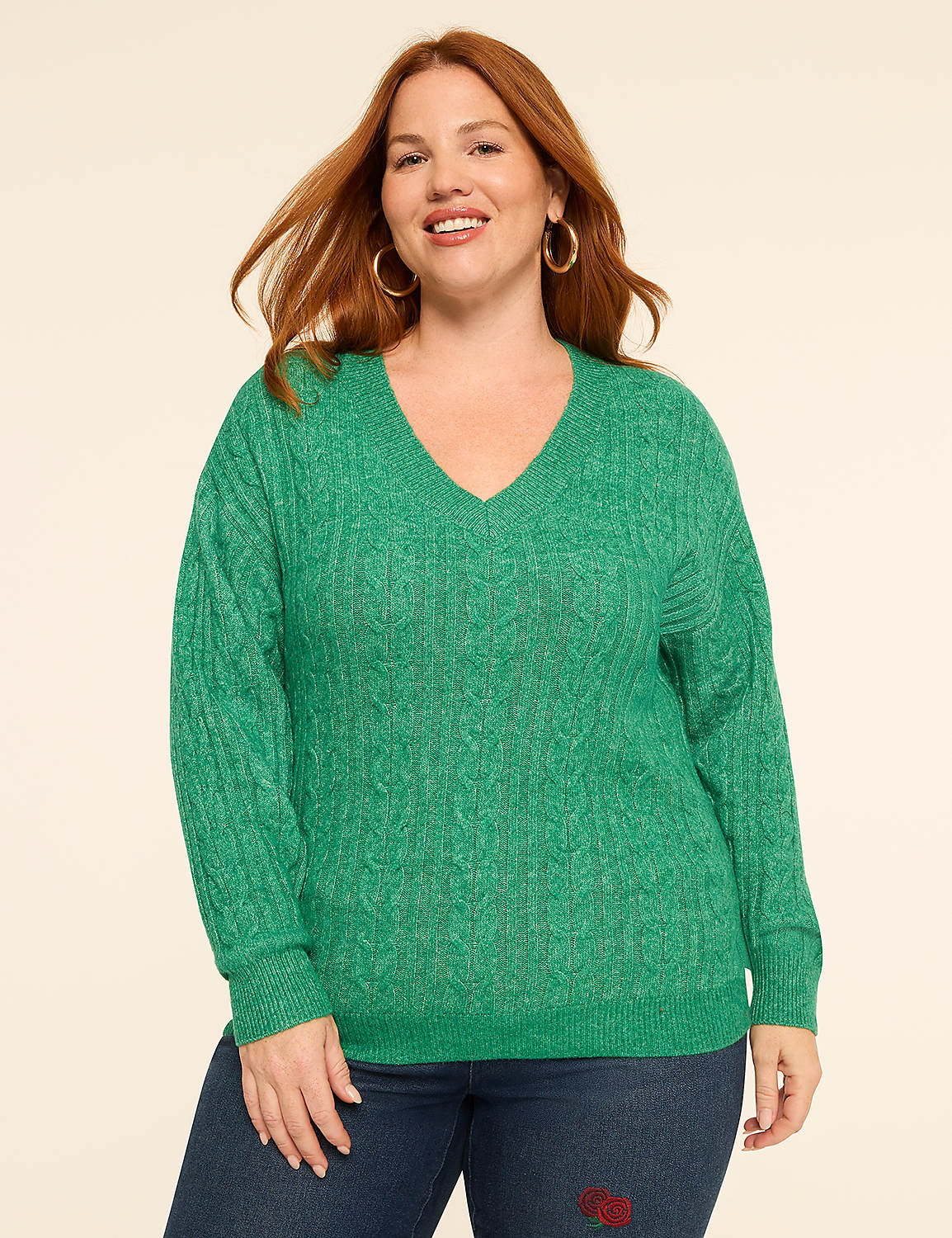 A woman with red hair smiles in a green cable-knit sweater and blue jeans against a plain, light-colored background. Product Image 1