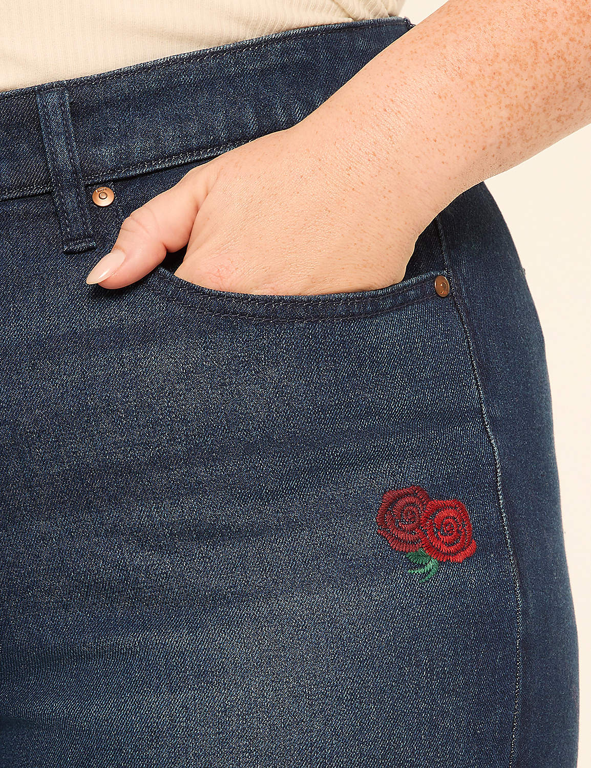 A hand rests in the pocket of dark wash jeans embroidered with two red roses, against a plain cream background. Product Image 4