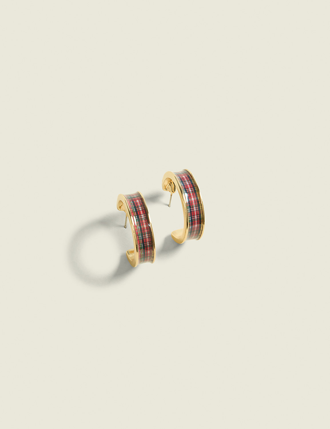 A pair of gold hoop earrings with a red, green, and black plaid pattern rest on a plain, light beige background. Product Image 1