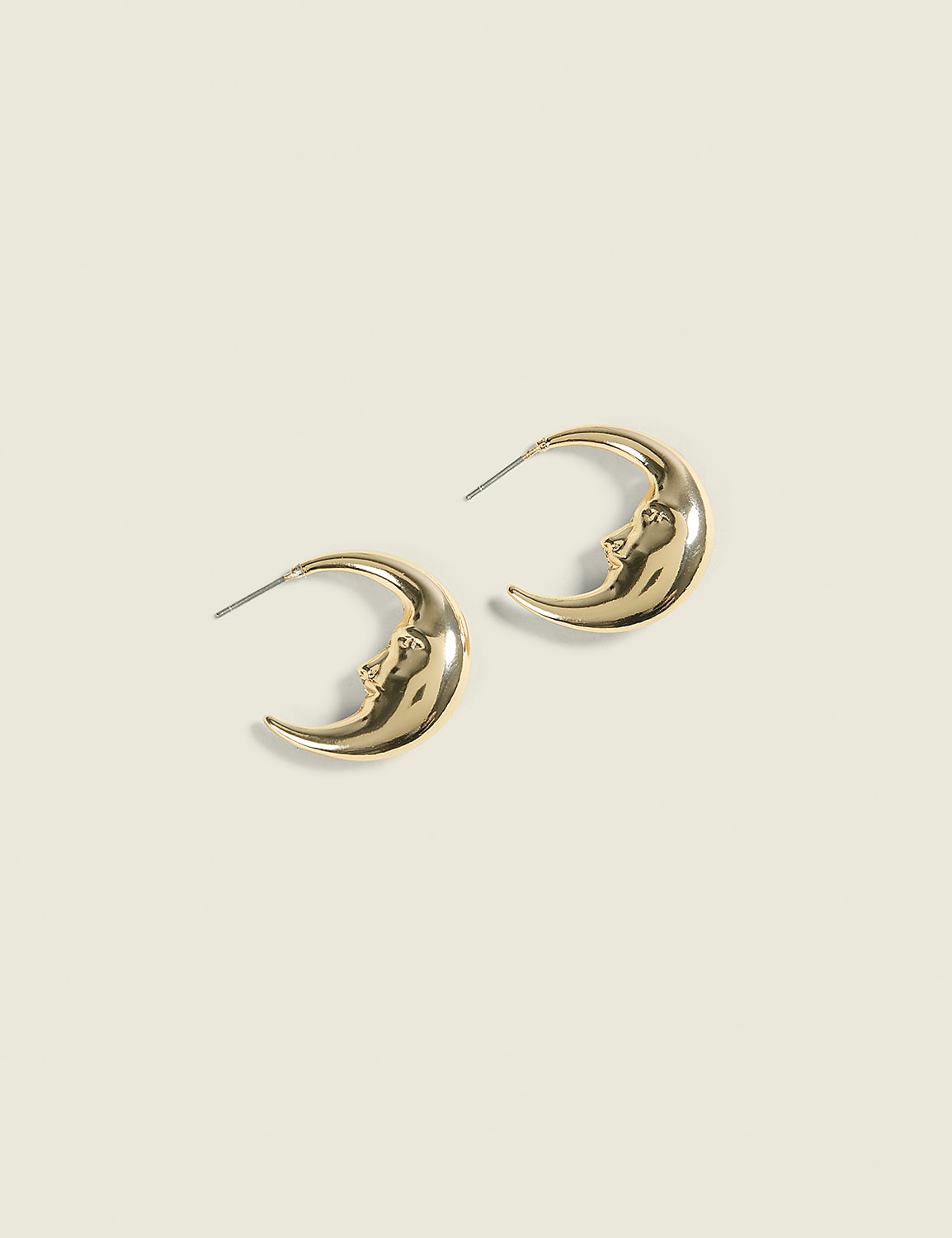 A pair of golden crescent moon earrings with faces are laid out on a pale, textured background. Product Image 1