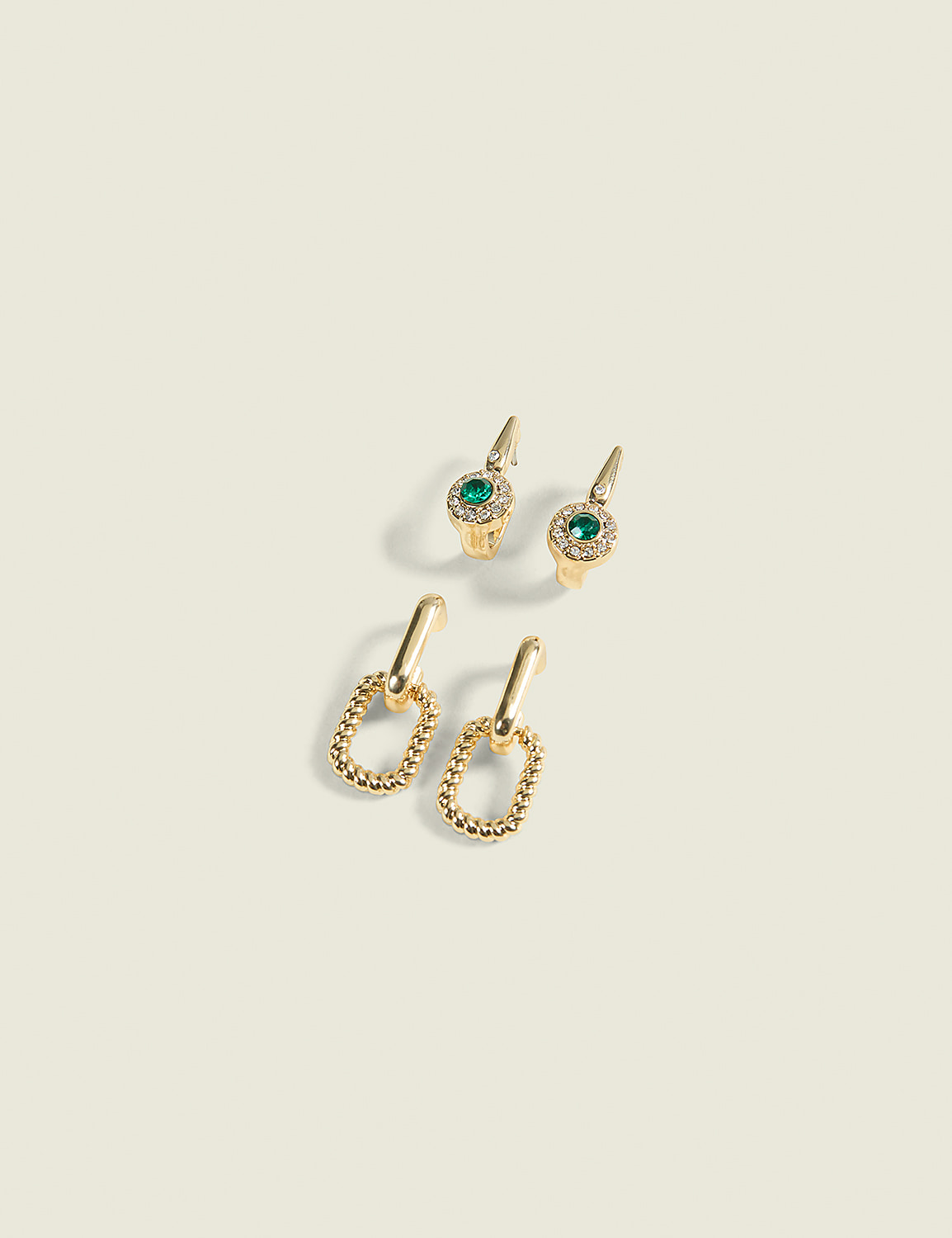 Two pairs of gold earrings, one with emeralds and diamonds and the other with textured oval links, rest on a pale yellow background. Product Image 1
