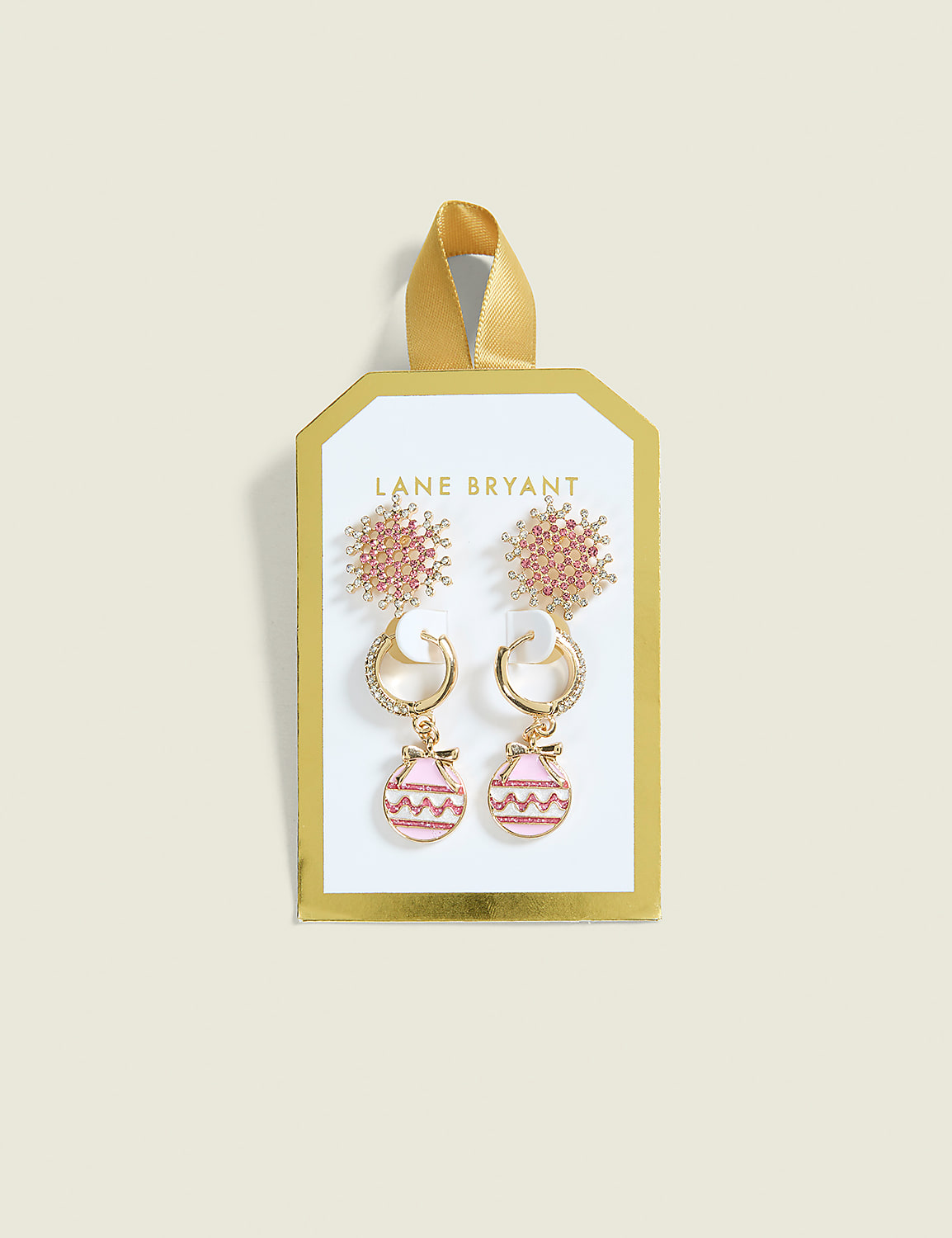 Two pairs of pink and gold earrings, one with a snowflake design and the other with ornament-shaped drops, are displayed on a white and gold card against a pale yellow background. Product Image 1