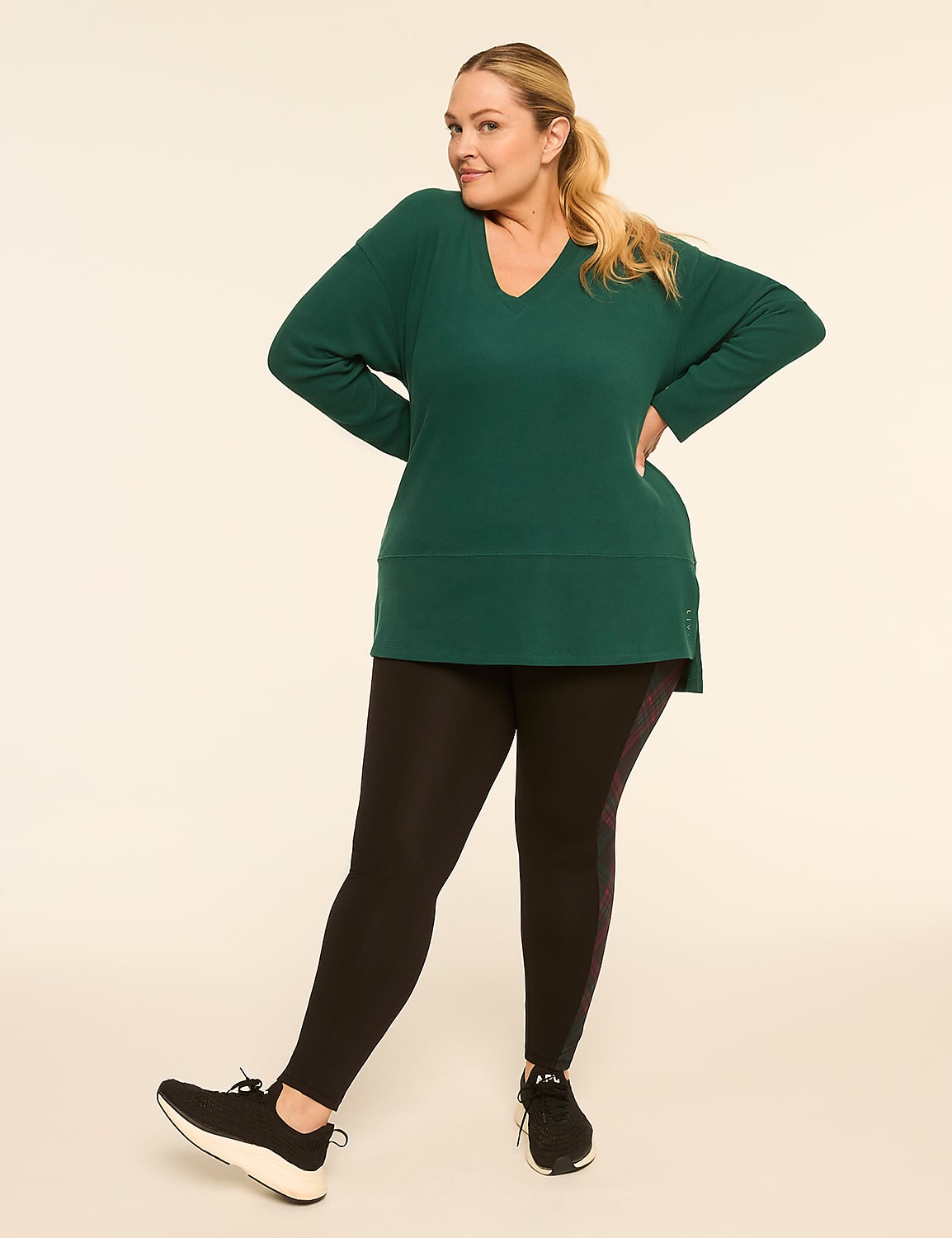 A woman in a green tunic and black leggings stands with her hands on her hips against a plain, light-colored background. Product Image 3