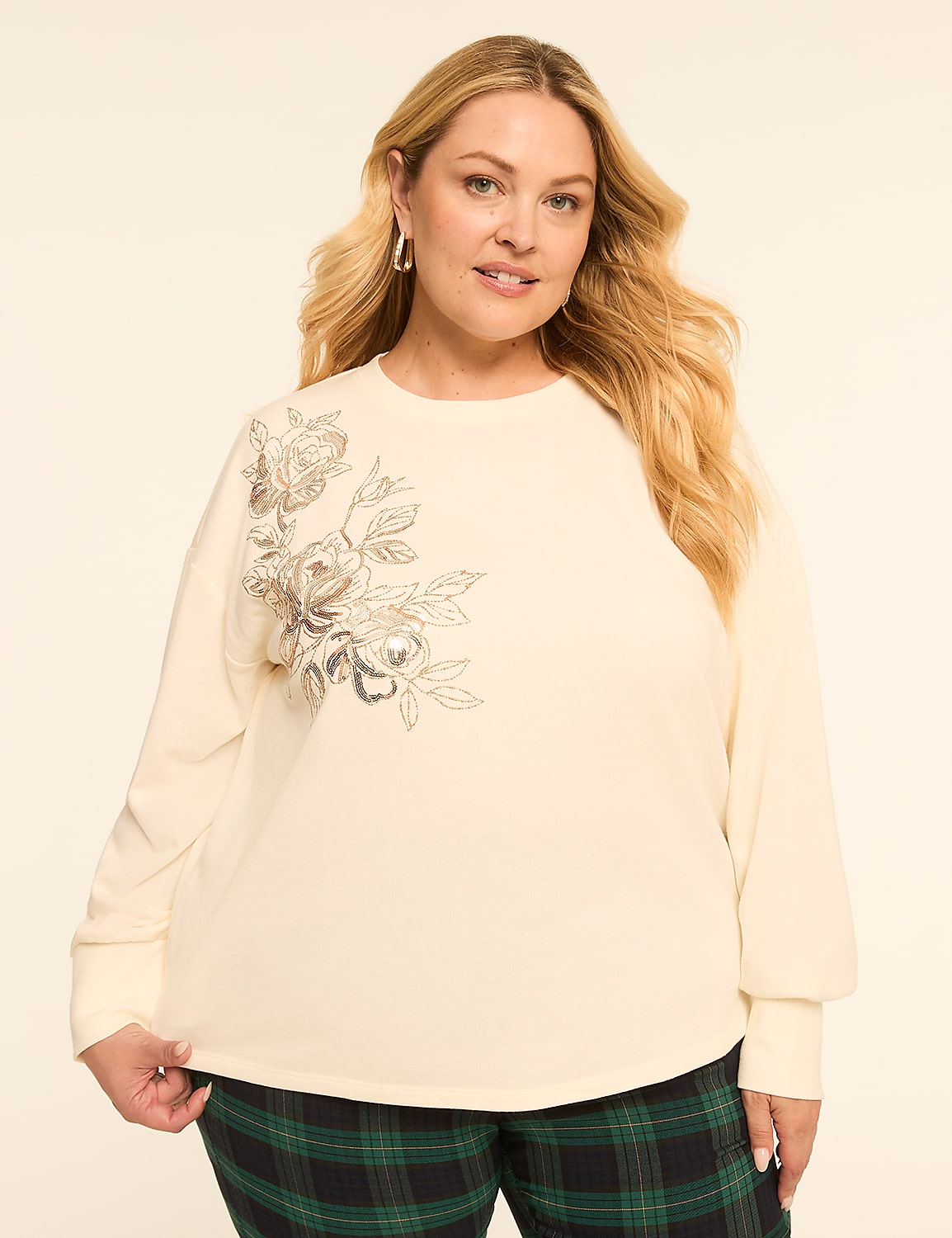 A woman with blonde hair and a cream-colored sweatshirt with floral embellishments stands against a plain, light-colored background, wearing plaid pants. Product Image 1