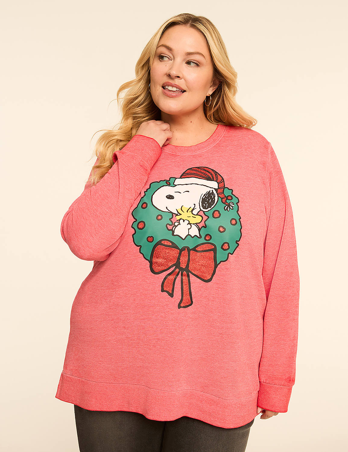 A woman in a coral sweatshirt with a Snoopy Christmas wreath design stands against a plain, light-colored background. Product Image 1