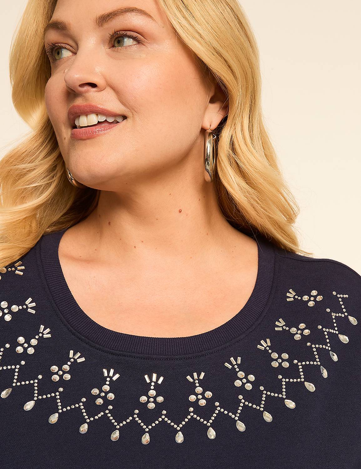 A woman with blonde hair and hoop earrings smiles as she wears a navy blue top adorned with silver embellishments, against a plain beige background. Product Image 4