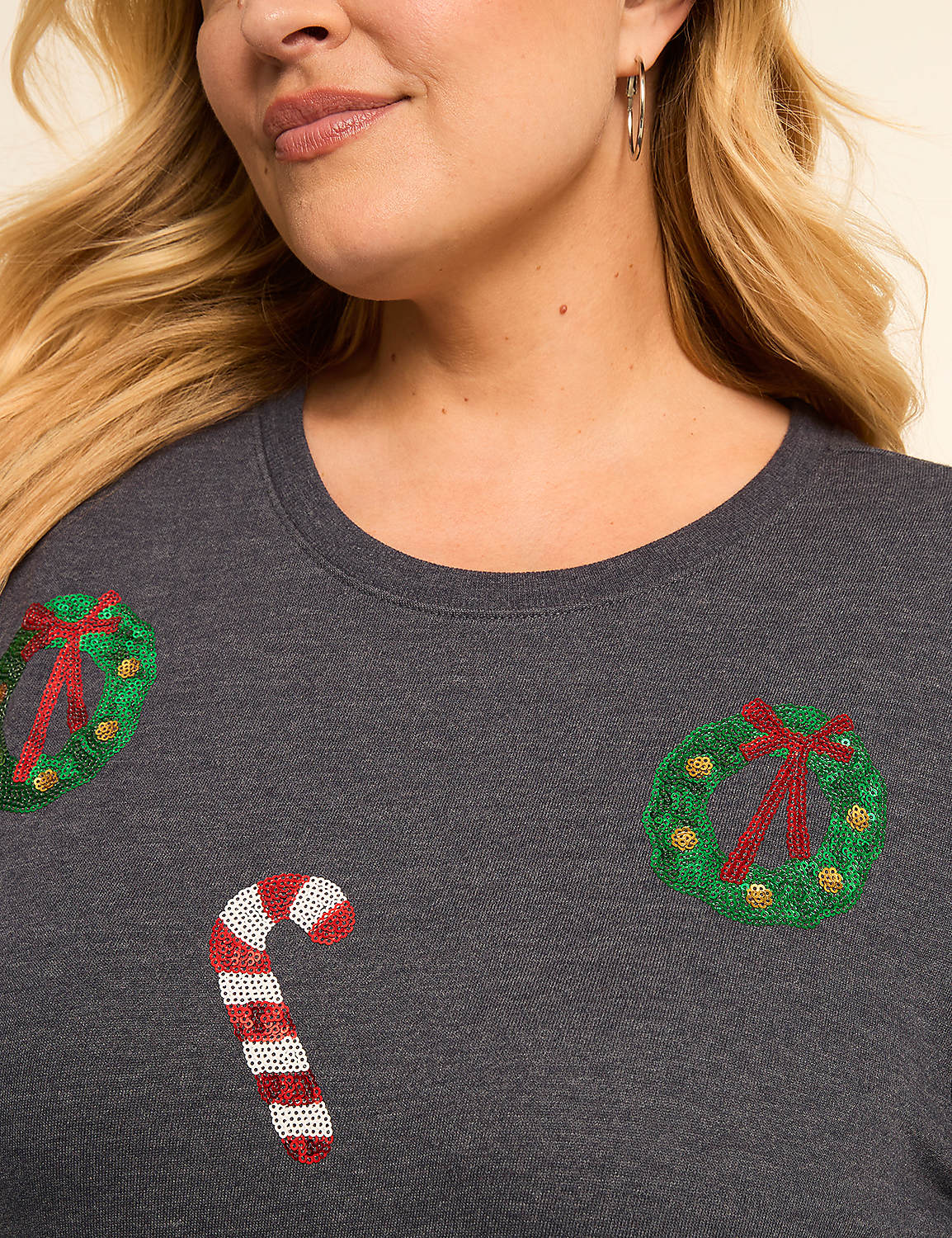 A woman with blonde hair and hoop earrings smiles as her dark gray t-shirt, adorned with sequined Christmas wreaths and a candy cane, is shown in the foreground against a plain background. Product Image 4