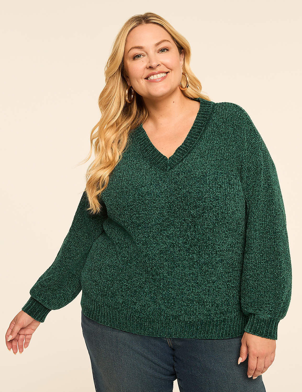 A smiling woman with blonde hair wears a green v-neck sweater and blue jeans against a plain, light-colored background. Product Image 1