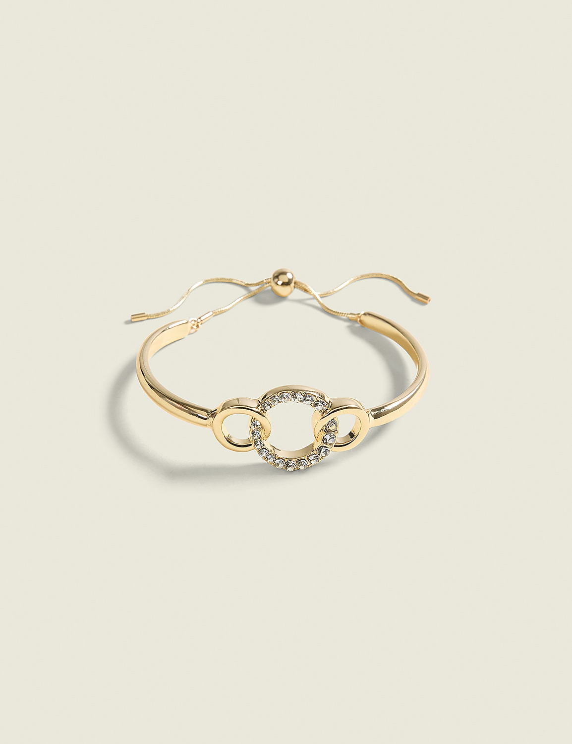 A gold bracelet with a circular, rhinestone-embellished charm and two smaller circular links rests on a pale green background. Product Image 1
