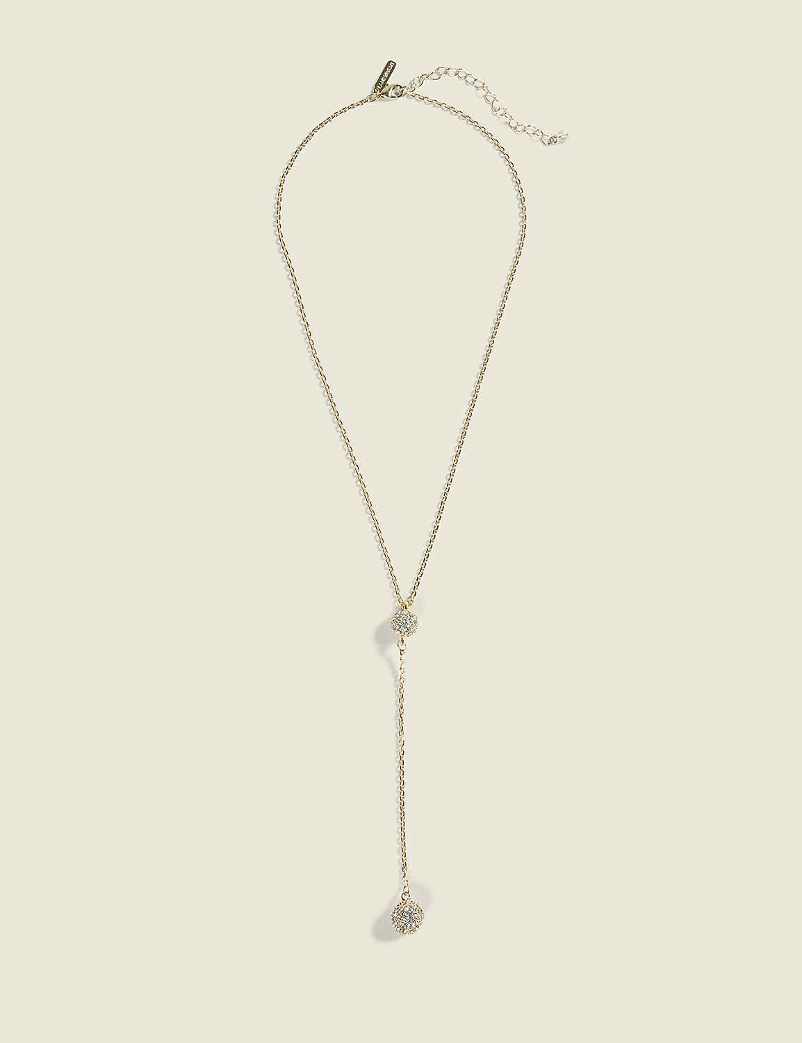 A delicate gold lariat necklace with a sparkling pendant hangs against a plain, light beige background. Product Image 1