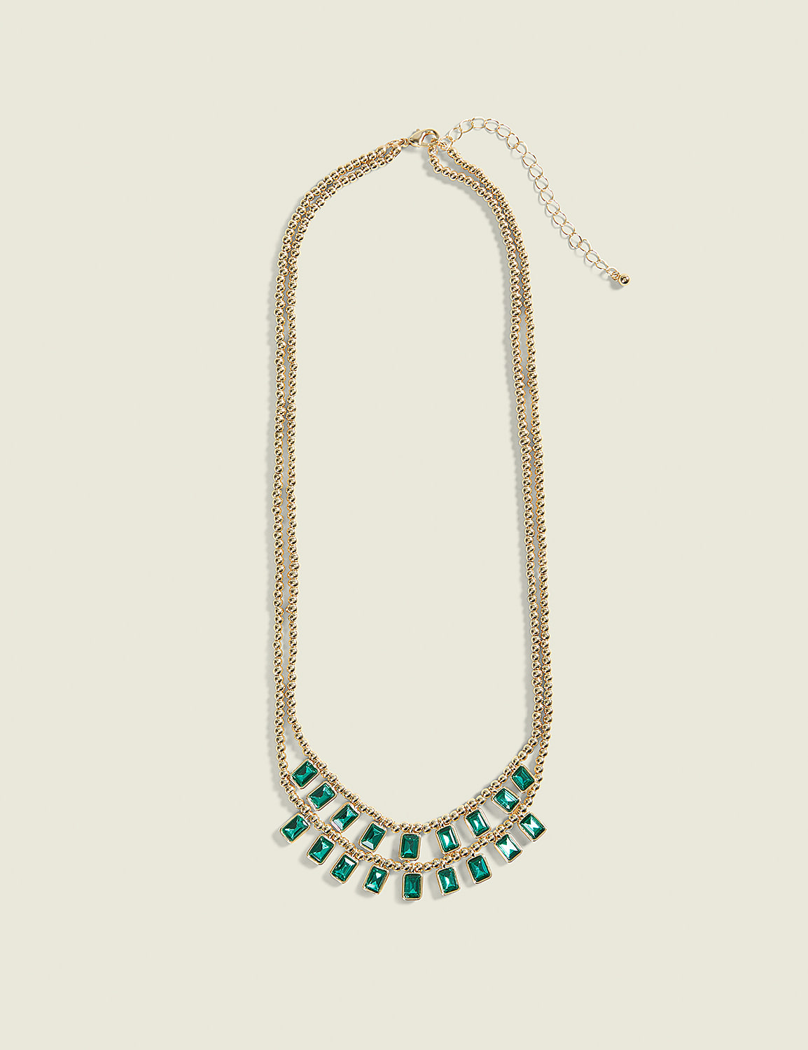 A gold necklace with emerald-colored rectangular stones hangs against a plain cream background. Product Image 1
