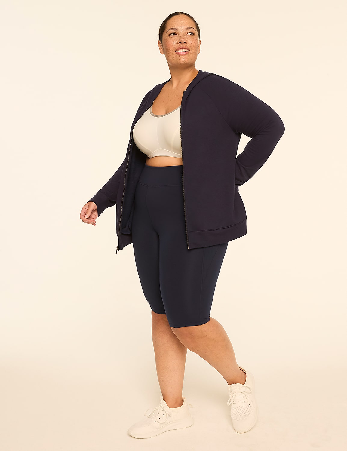 A woman in a navy zip-up hoodie, cream sports bra, and navy bike shorts walks against a plain, light-colored background. Product Image 3