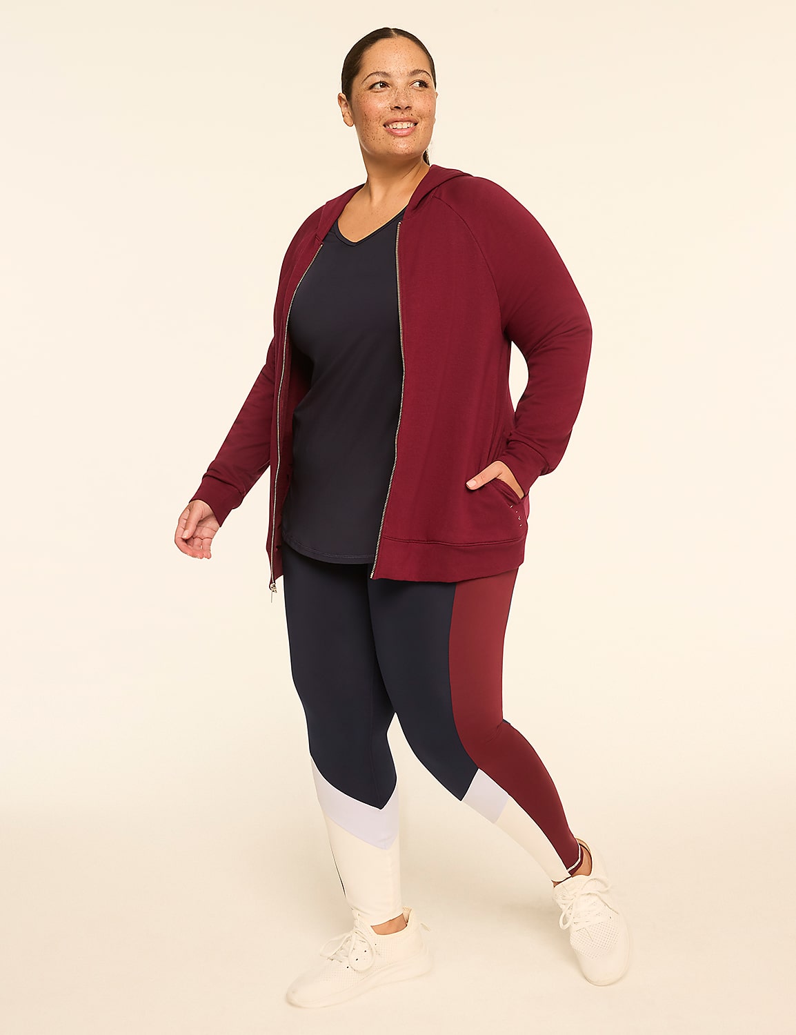 A woman in a maroon zip-up hoodie and color-blocked leggings walks against a plain, light-colored background. Product Image 3