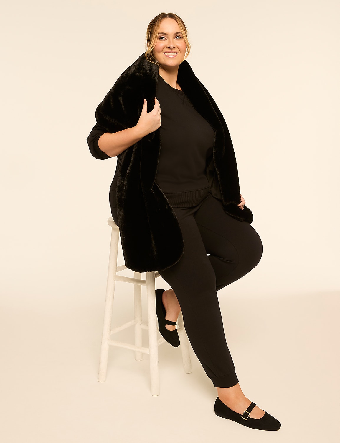 A smiling woman in a black outfit and faux fur stole sits on a white stool against a cream-colored background. Product Image 4