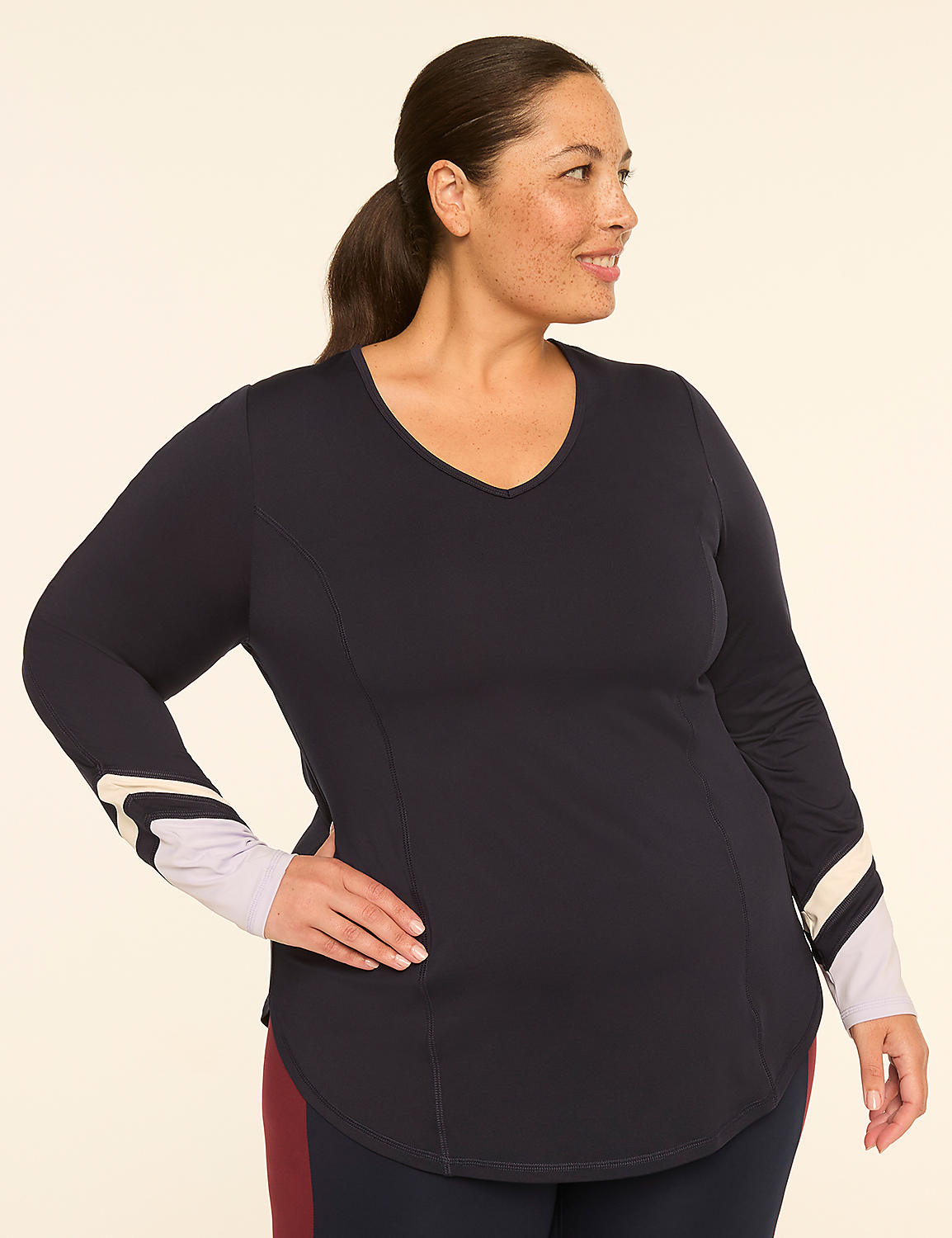 A woman with dark hair pulled back in a ponytail poses with her hand on her hip, wearing a navy v-neck shirt with striped cuffs, against a plain, light-colored background. Product Image 1