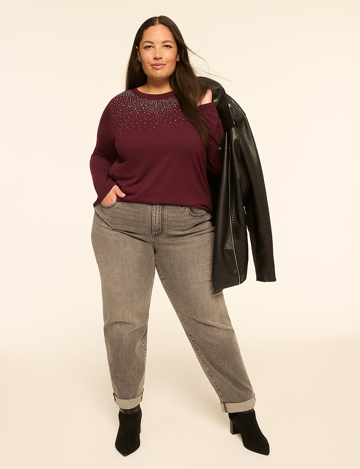 A woman in a burgundy top and grey jeans stands against a plain, light background, holding a black leather jacket. Product Image 3
