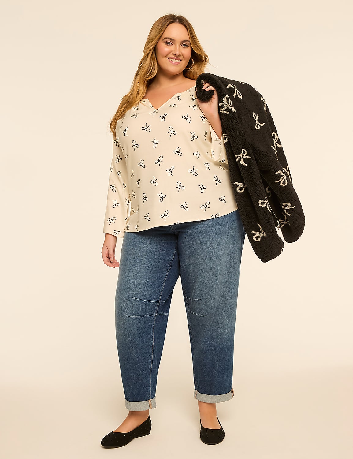 A smiling woman in a cream-colored top with a bow pattern and blue jeans stands against a plain, light-colored background, holding a black jacket with a bow pattern over her shoulder. Product Image 3