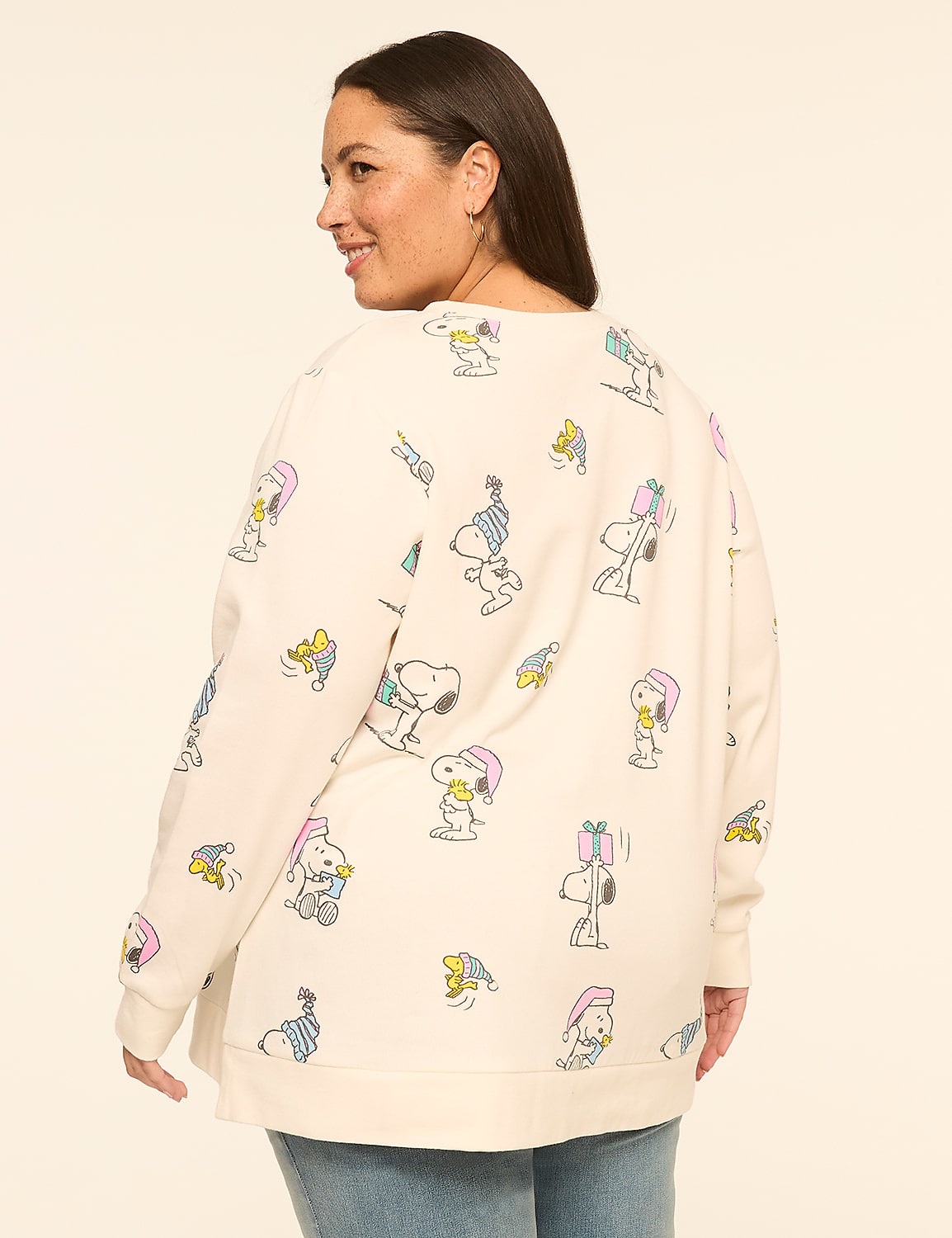 A woman wearing a cream-colored sweatshirt with a Snoopy holiday print stands against a plain, light-colored background. Product Image 2