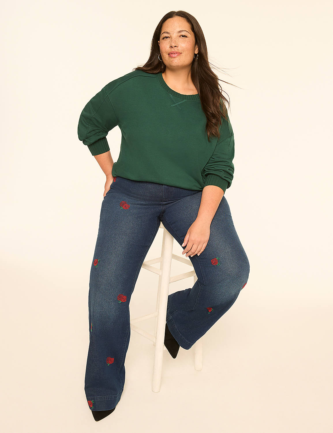 A woman with dark hair and a warm smile sits on a white stool in the foreground, wearing a green sweatshirt and embroidered jeans, against a plain, light-colored background. Product Image 4