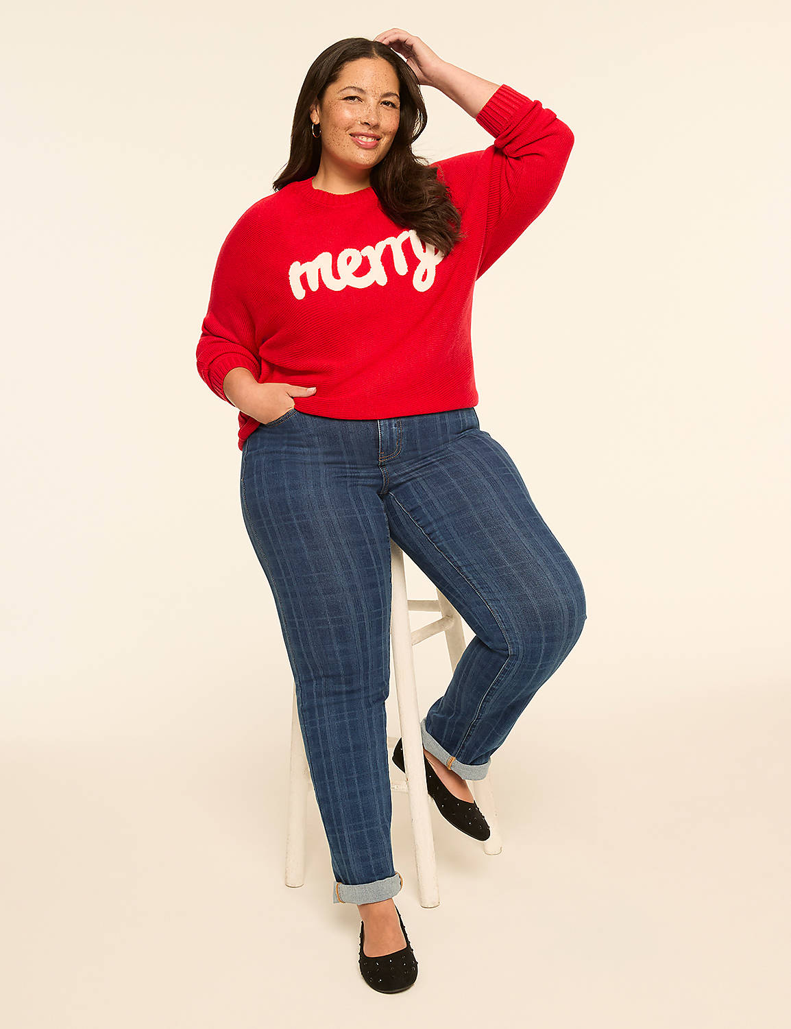 A woman in a red "merry" sweater and striped jeans sits on a stool against a plain, light-colored background. Product Image 3