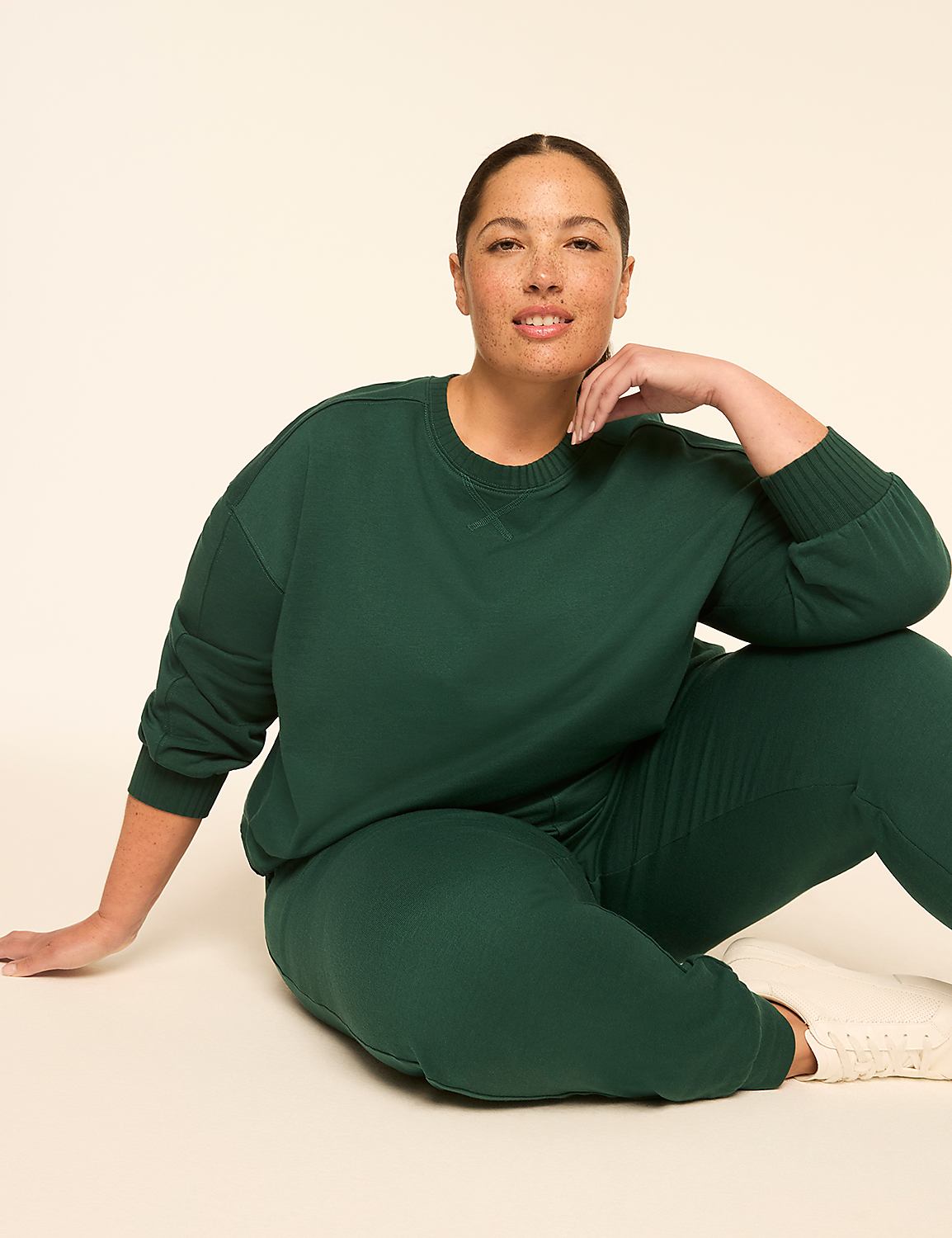 A woman in a green sweatshirt and sweatpants sits on a light-colored floor with a plain, light background. Product Image 3