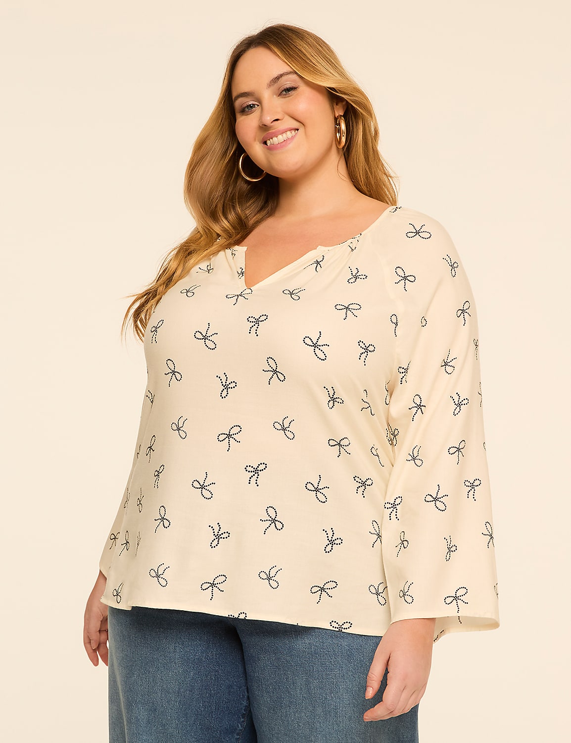 A smiling woman in a cream-colored top with a bow pattern and blue jeans stands against a plain beige background. Product Image 1