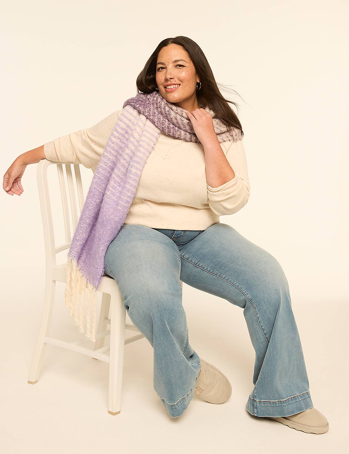 A woman in a cream sweater, light wash jeans, and a purple and cream scarf sits on a white chair against a plain, light background. Product Image 3