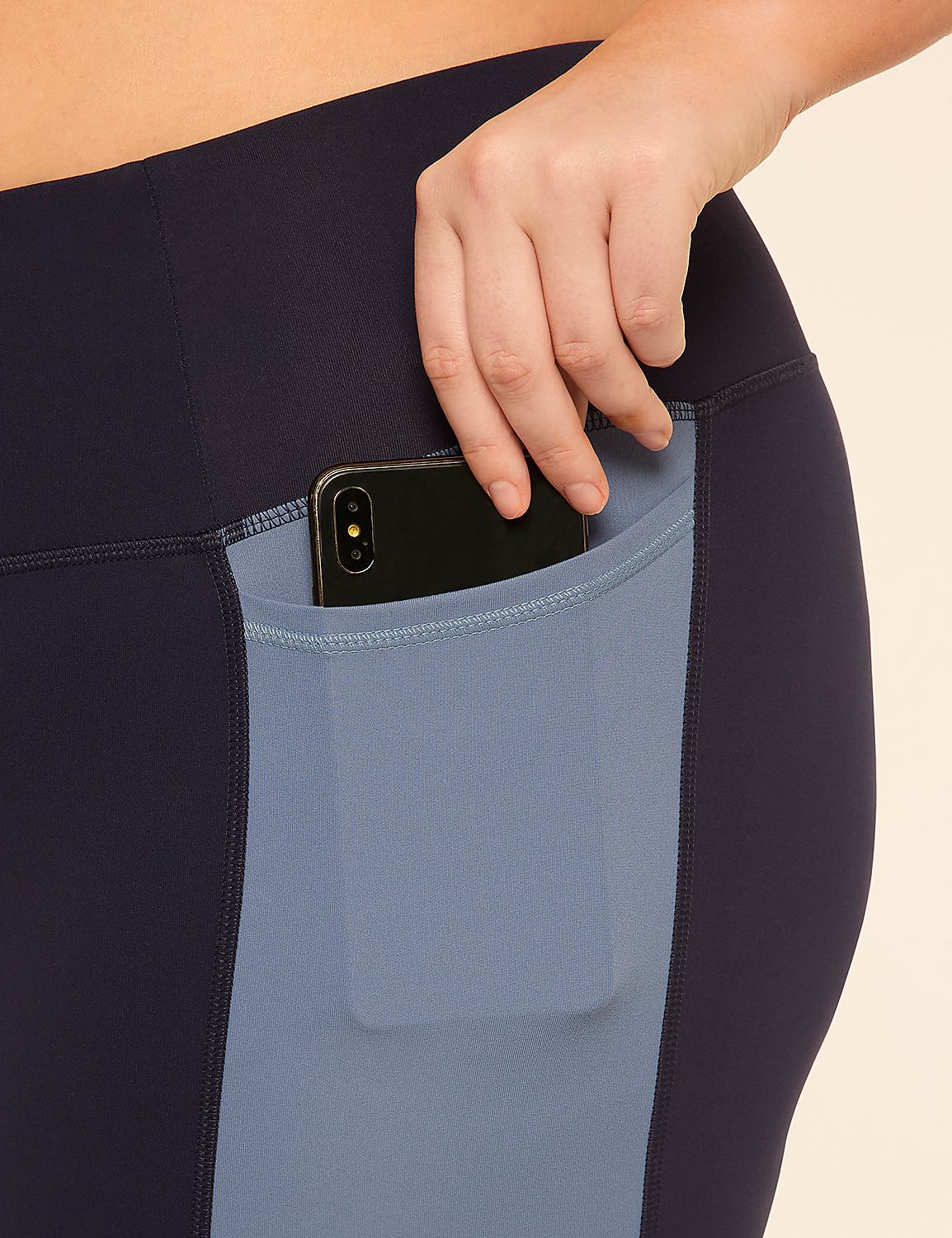 A hand places a smartphone into the side pocket of navy and blue athletic leggings against a plain background. Product Image 4