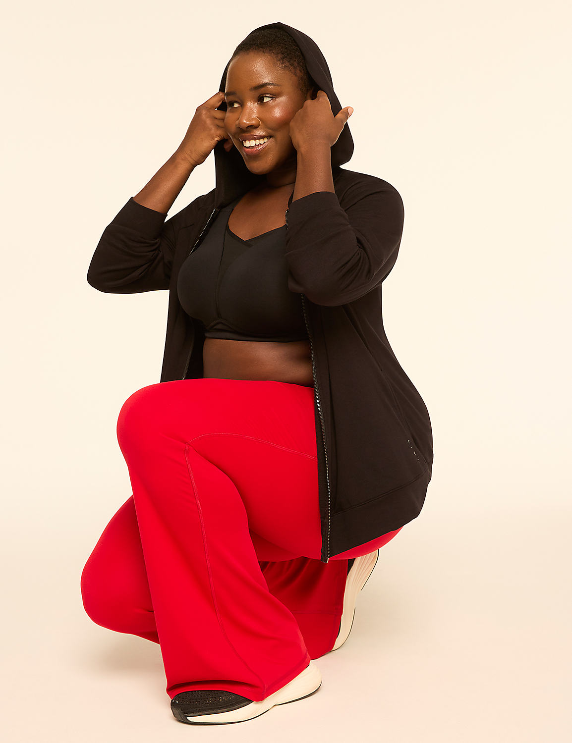 A woman in a black hoodie and red pants smiles while adjusting her hood against a plain, light-colored background. Product Image 3