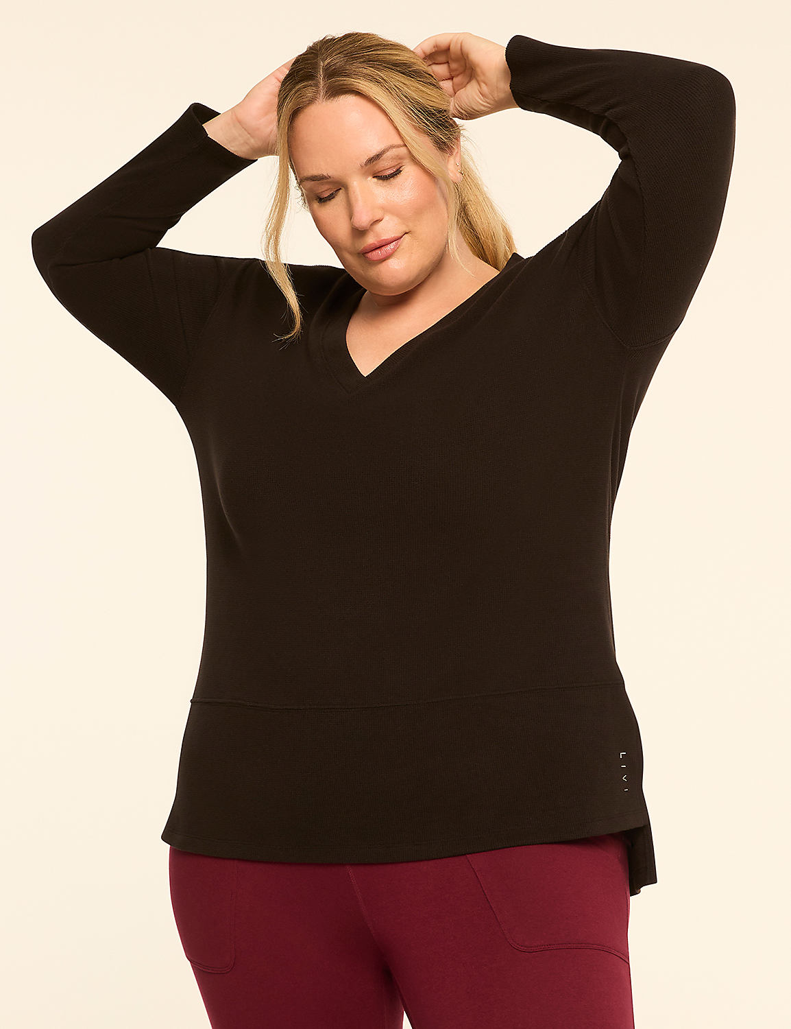 A woman in a black v-neck top and maroon leggings poses with her hands behind her head against a plain cream background. Product Image 1