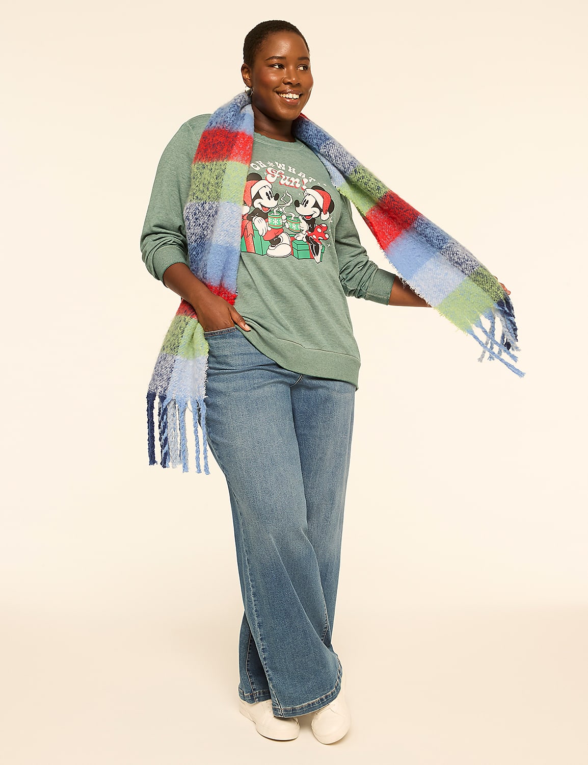 A woman in a green Mickey Mouse sweatshirt and wide-leg jeans stands against a plain, light background, draped in a colorful plaid scarf. Product Image 3