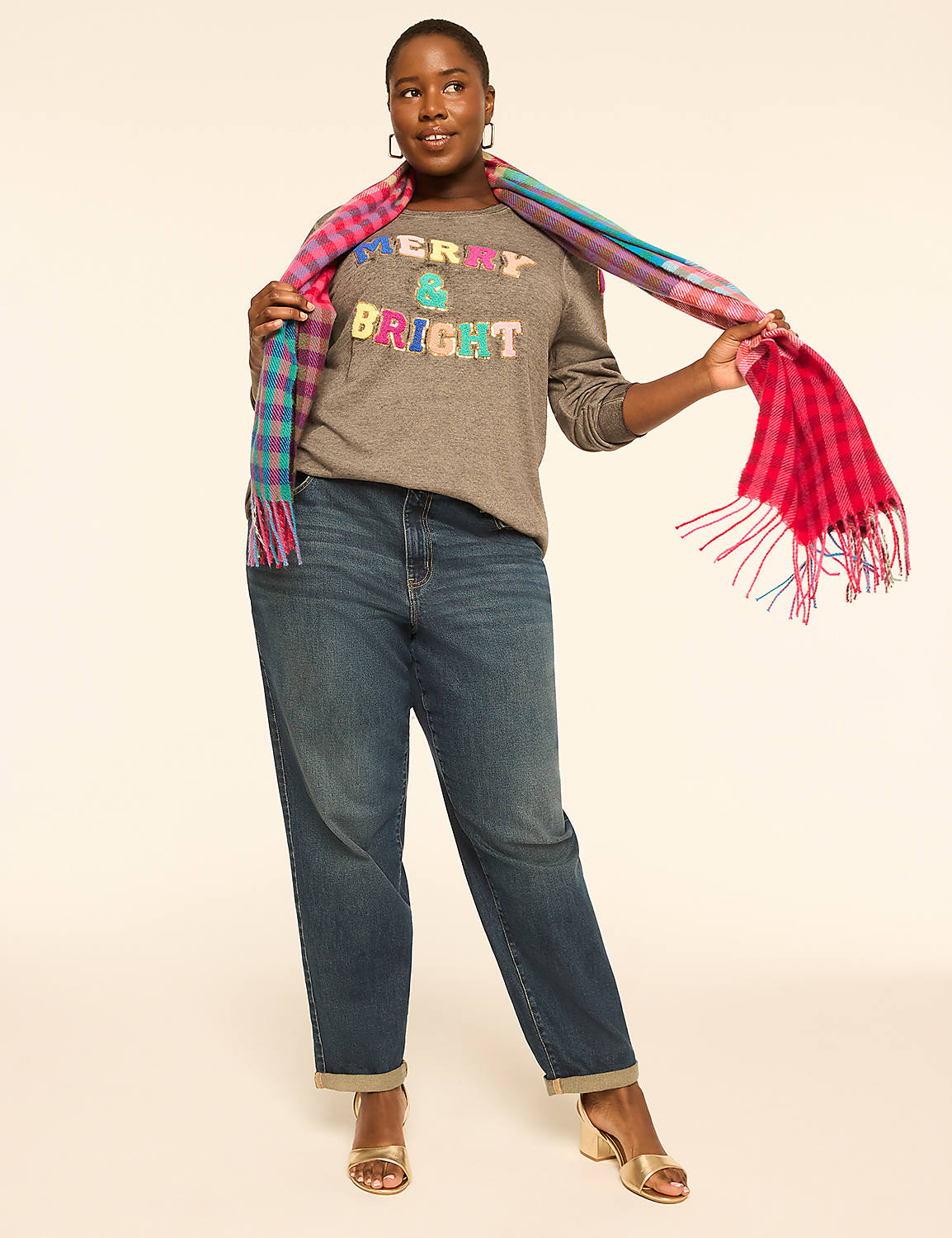 A woman in a gray "Merry & Bright" sweatshirt and blue jeans poses with a colorful plaid scarf against a plain, light-colored background. Product Image 3