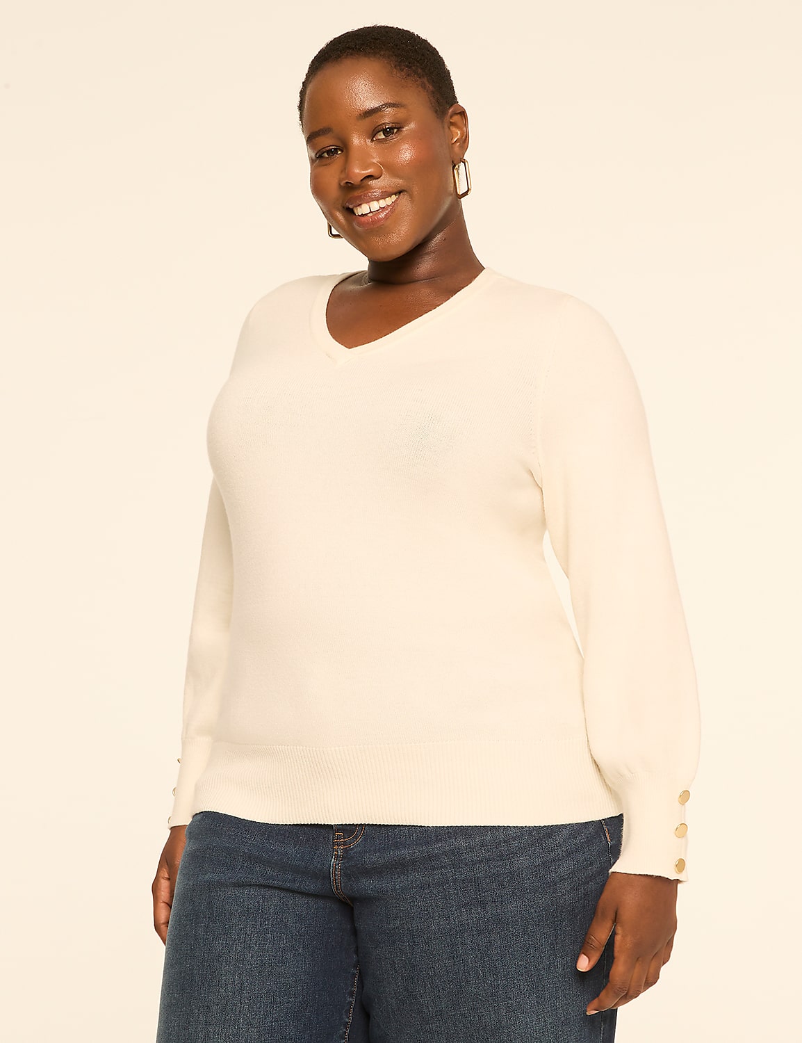 A smiling woman in a cream v-neck sweater and blue jeans stands against a plain, light-colored background. Product Image 1