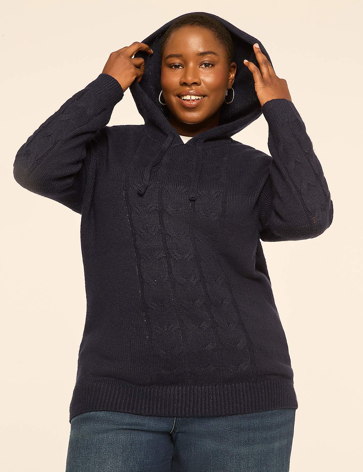 A woman in a navy cable-knit hoodie and jeans smiles as she pulls the hood up against a plain, light-colored background. Product Image 1