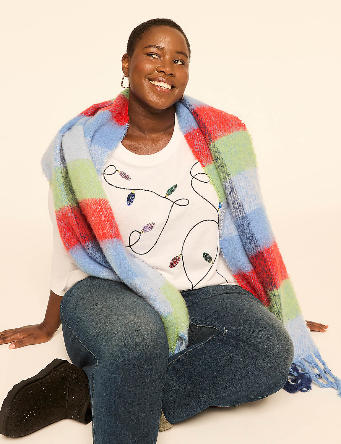 A woman in a white t-shirt with a string of Christmas lights design and blue jeans is wrapped in a colorful plaid scarf, sitting against a plain, light-colored background. Product Image 3