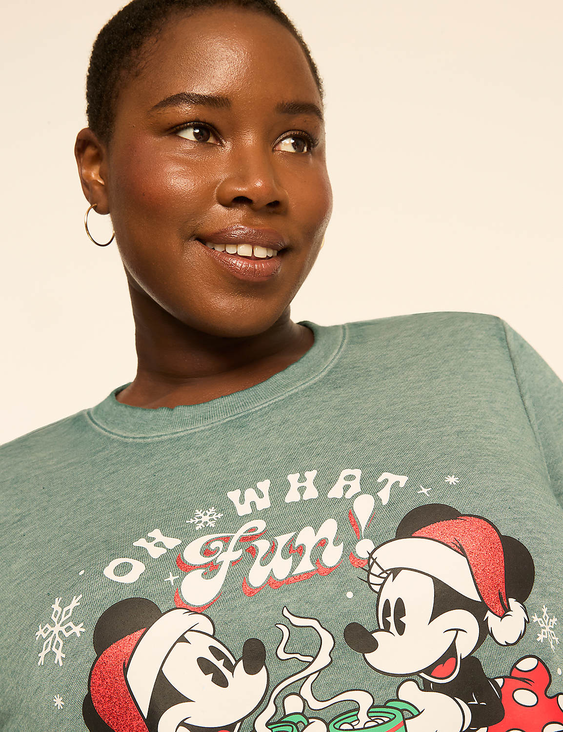 A woman with short hair smiles as she wears a green sweatshirt with Mickey and Minnie Mouse in Santa hats, with "Oh What Fun!" printed above them. Product Image 4