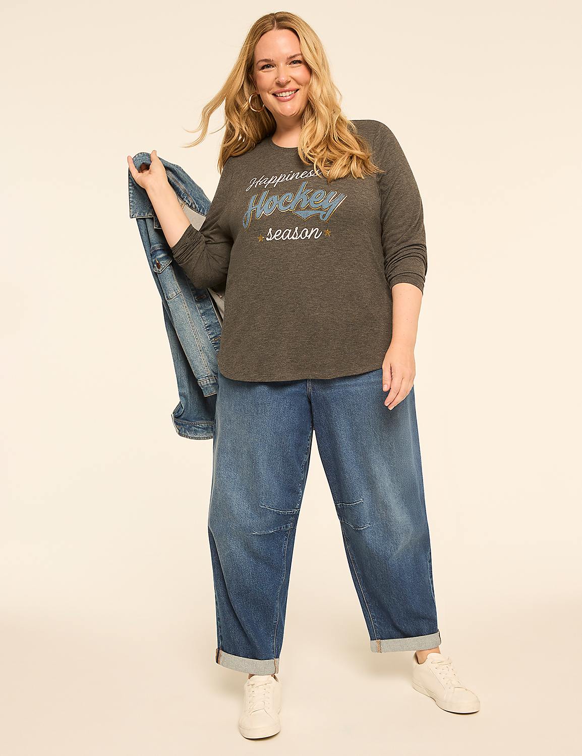 A smiling woman in a grey t-shirt and baggy jeans stands against a plain, light-colored background, holding a denim jacket. Product Image 3