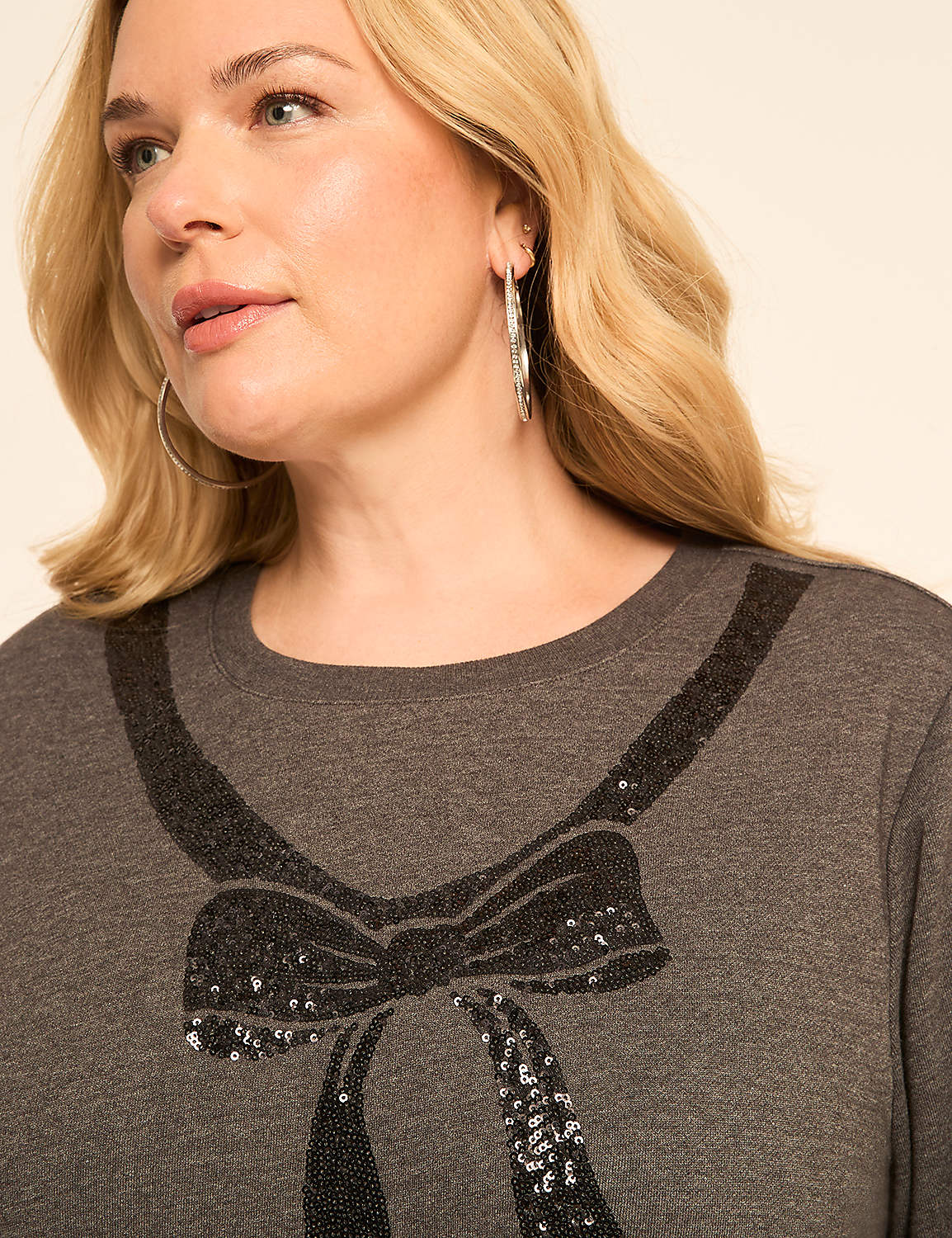 A woman with blonde hair and hoop earrings wears a dark gray t-shirt with a sequined black bow design, set against a plain, light background. Product Image 4