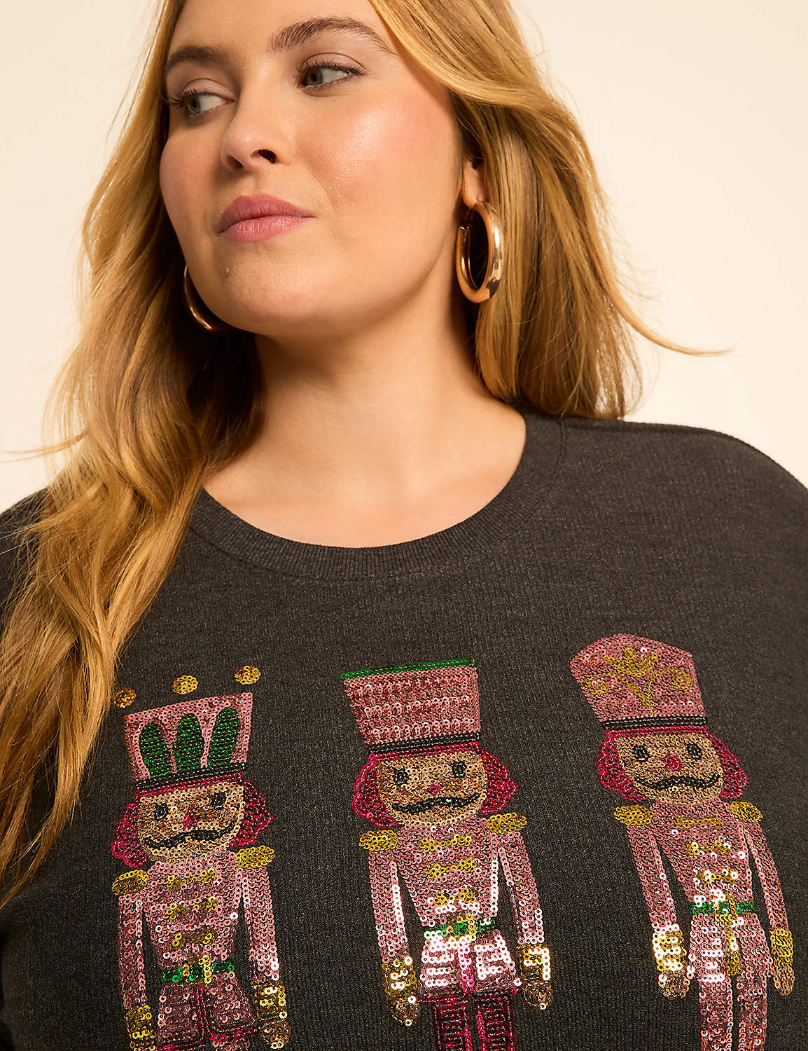 A woman with blonde hair and hoop earrings wears a dark gray sweater adorned with three sequined nutcracker figures in the foreground, against a plain, light-colored background. Product Image 4
