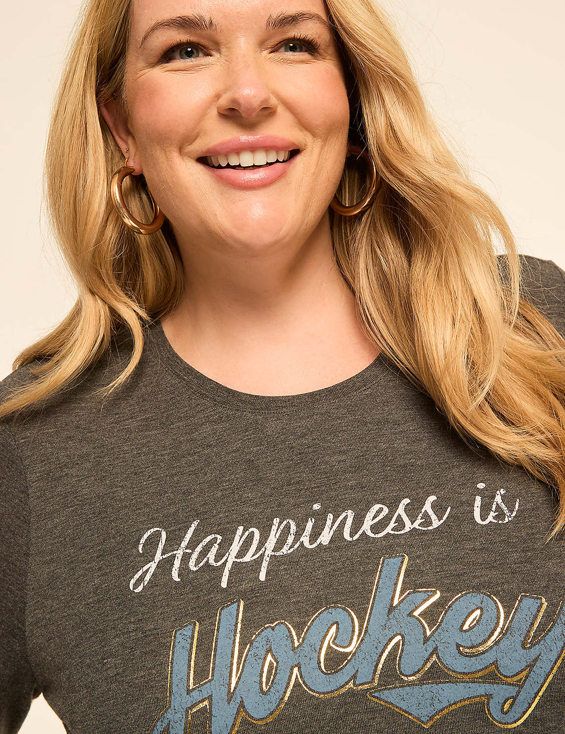 A smiling woman with blonde hair and hoop earrings wears a dark gray t-shirt with "Happiness is Hockey" printed on it, against a plain, light background. Product Image 4