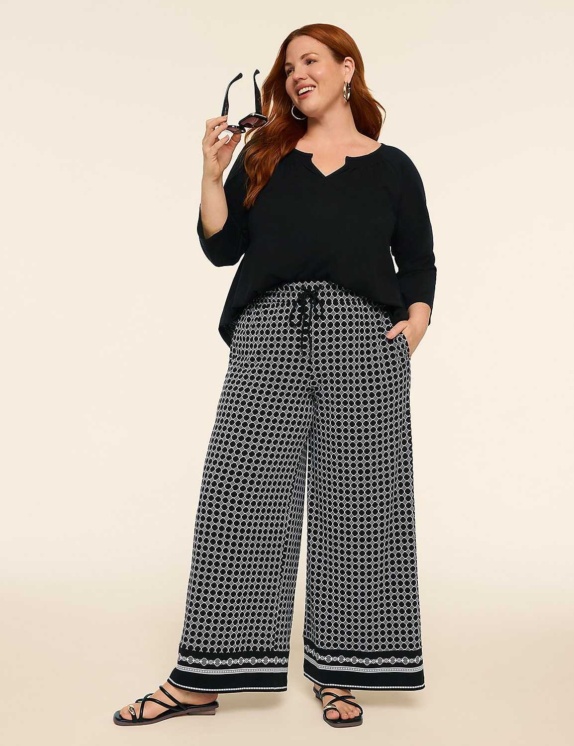 A woman with red hair stands in the foreground wearing a black top and patterned wide-leg pants, with a plain, light-colored background. Product Image 3