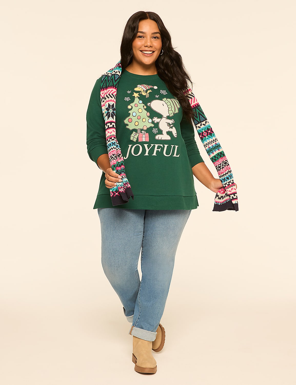 A smiling woman in a green "JOYFUL" Christmas sweater and light blue jeans stands against a plain, light tan background, with a colorful patterned scarf draped around her neck. Product Image 3